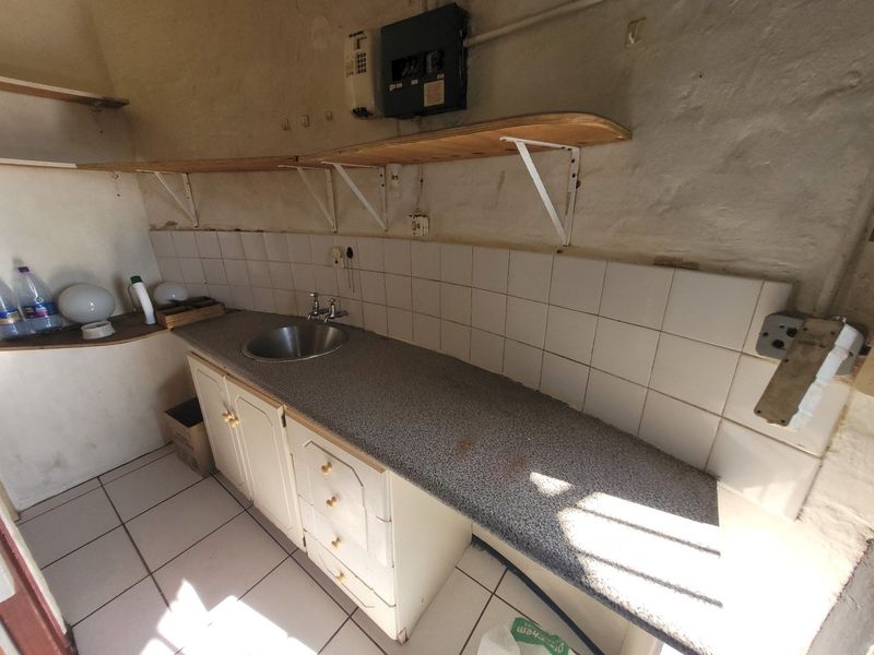 A functional scullery area featuring a stainless steel sink, tiled backsplash, built-in cabinetry, and wall-mounted shelving, requiring cosmetic updates.