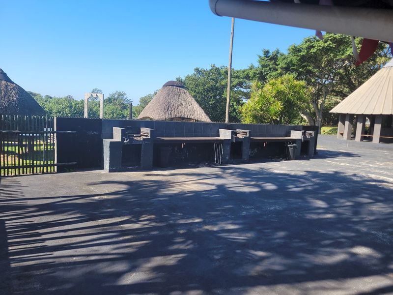 Outdoor entertainment area featuring a long built-in braai station with tiled splashback, set on a paved patio with surrounding thatched structures.