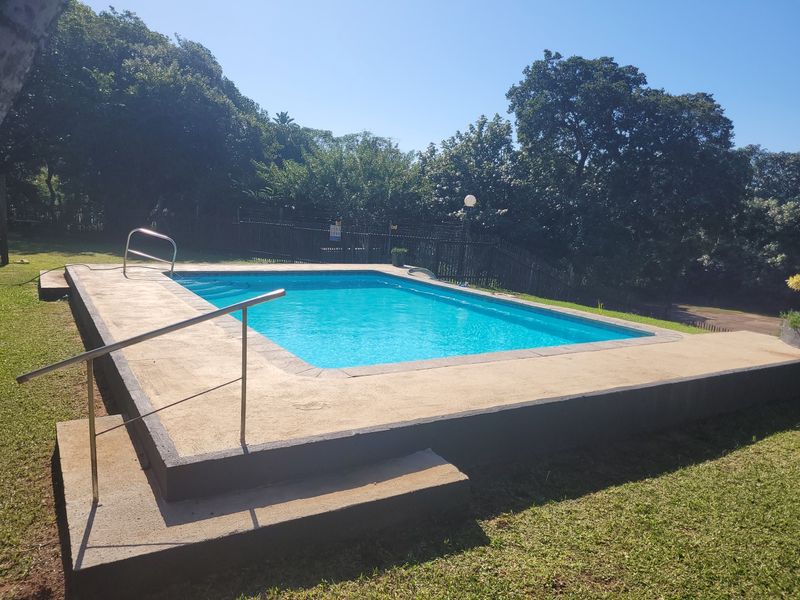 Outdoor swimming pool area featuring a concrete surround, metal safety handrails, and perimeter fencing set within a landscaped garden.