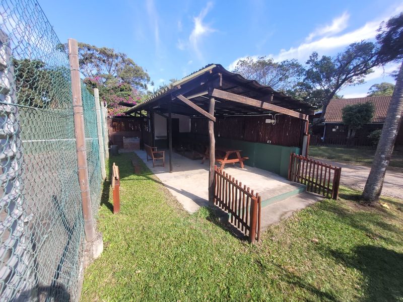 Outdoor covered patio area featuring a timber pergola structure, concrete flooring, and a built-in brick braai, set within a garden space.