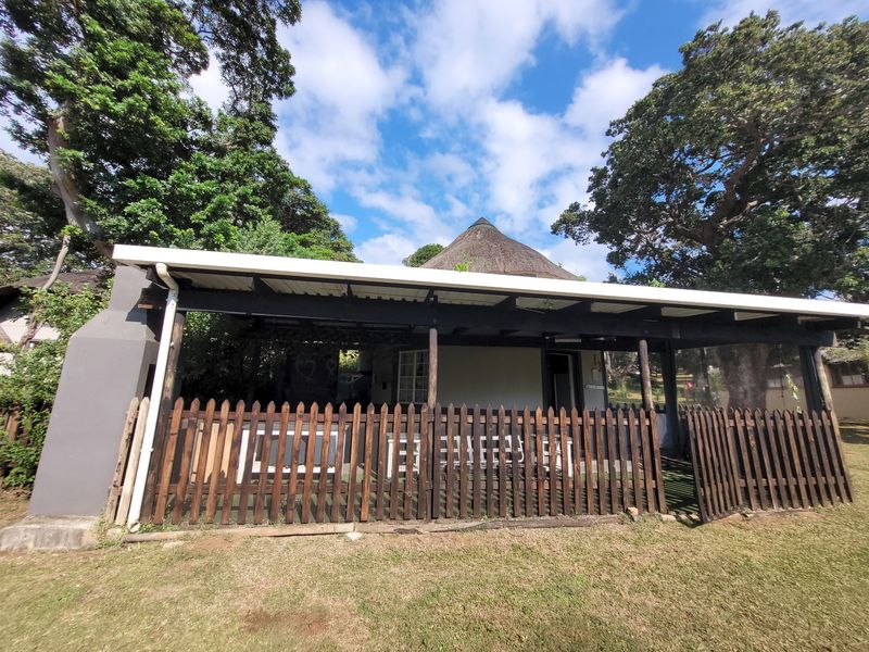 A covered outdoor patio area featuring a thatched roof, built-in fireplace, and timber picket fencing, set within a garden environment.