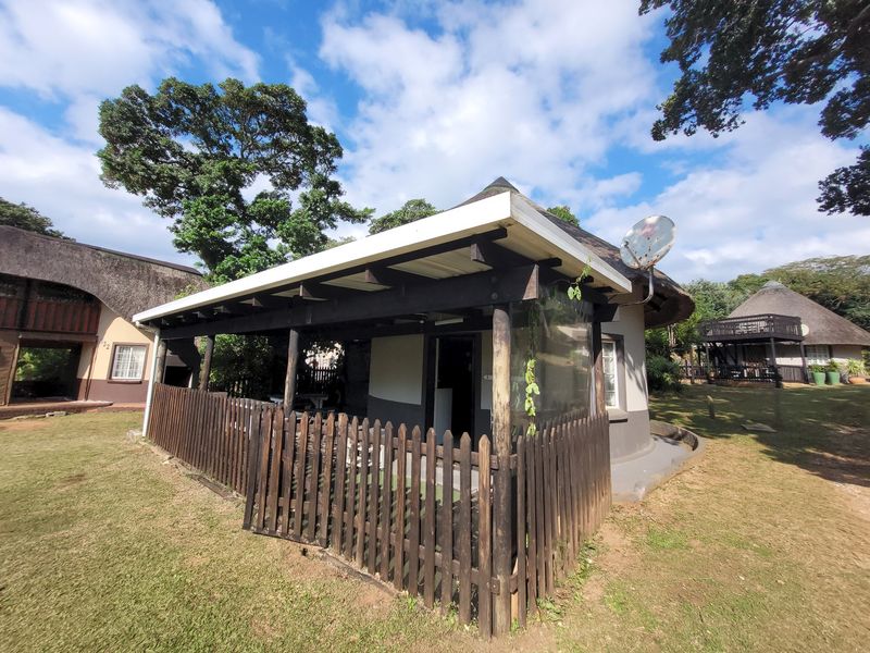 A covered patio area featuring timber support posts and a picket fence, set within a garden environment with a thatched roof structure.