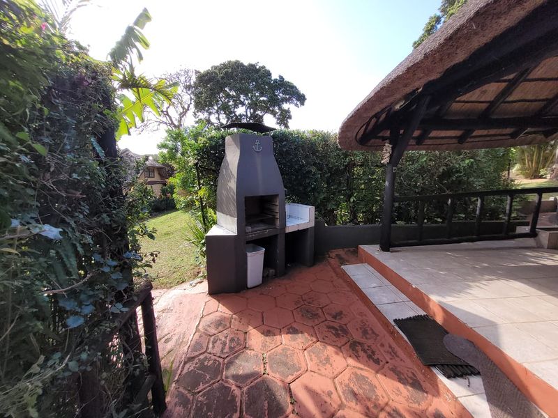 Outdoor patio area featuring a built-in braai, thatched roof structure, and paved flooring, set within a garden environment.