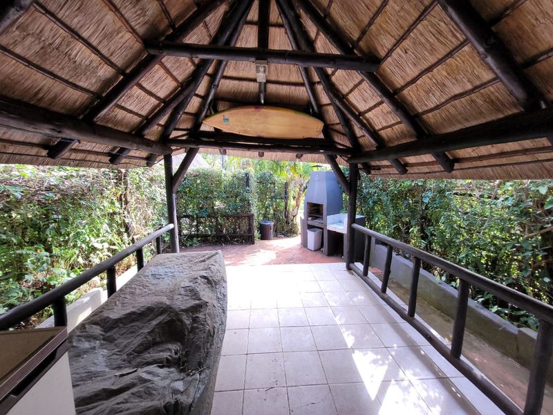 Covered outdoor patio featuring a traditional thatched roof, timber support structure, tiled flooring, and a built-in braai area, perfect for outdoor entertaining.