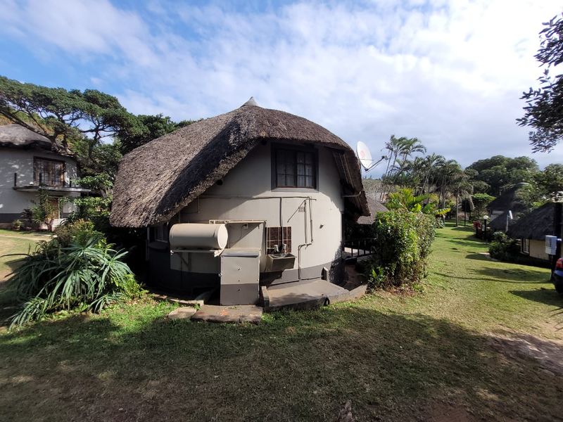 Rear view of a circular townhouse unit featuring a traditional thatch roof, external geyser, and wash basin set within a landscaped estate environment.