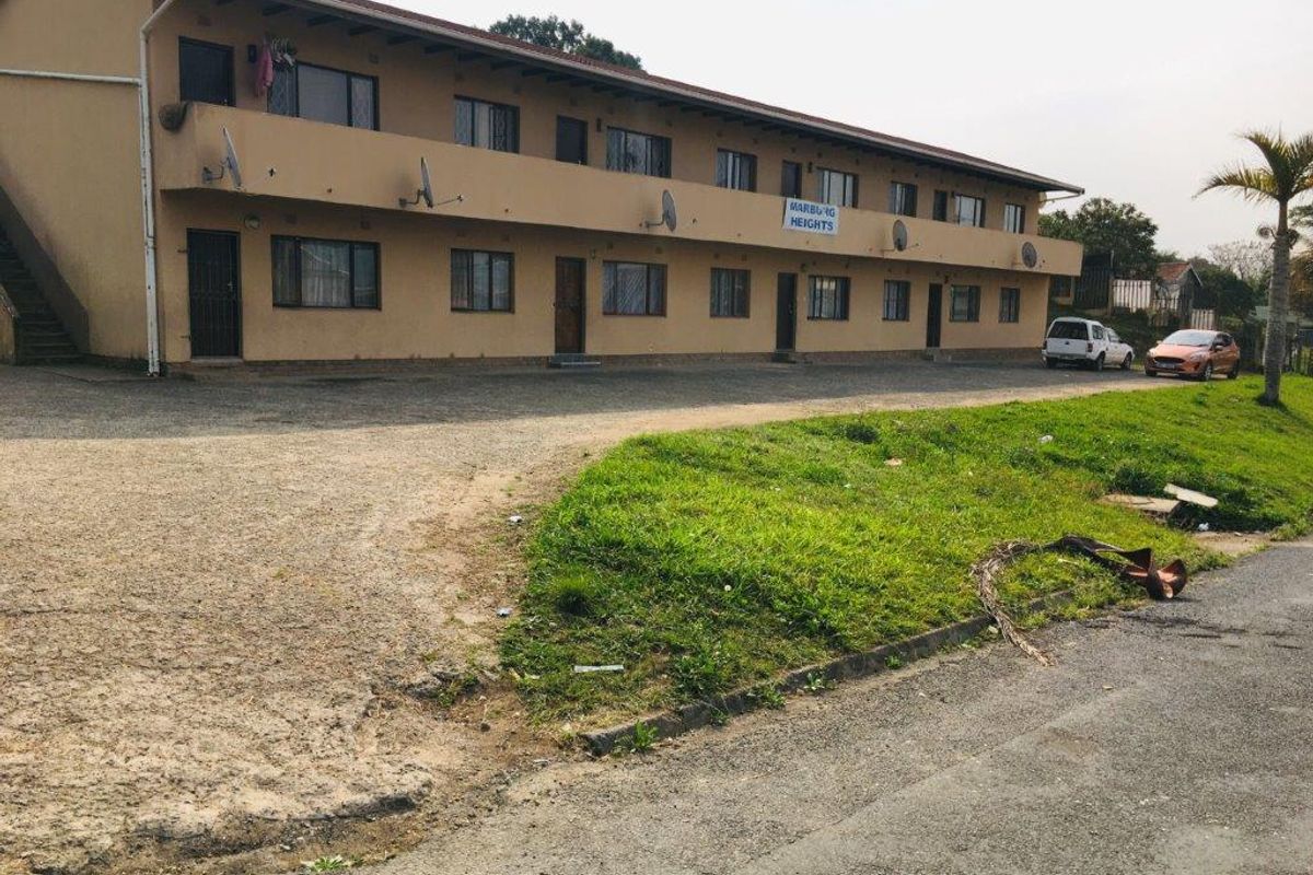 Exterior view of a multi-story apartment complex featuring a shared balcony, external staircase, and unpaved parking area.