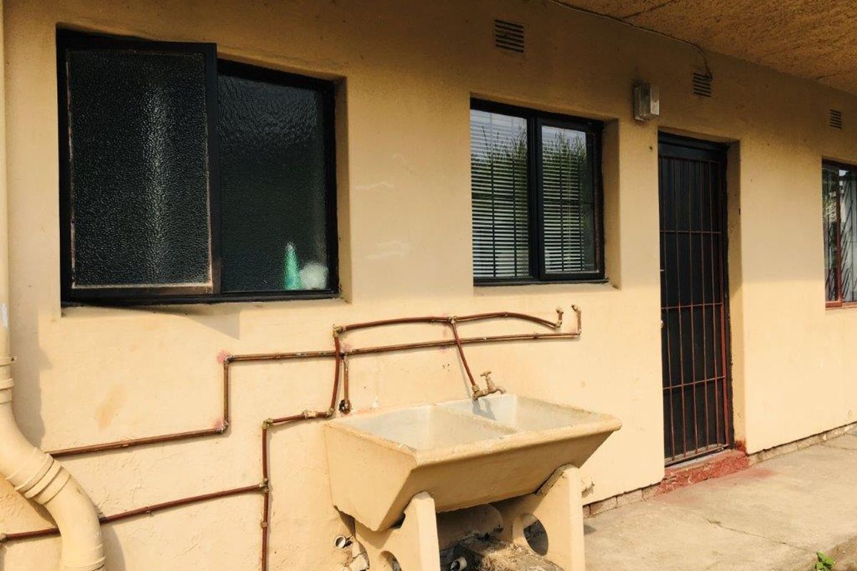 Rear exterior view of the apartment featuring a concrete laundry trough, exposed plumbing, and security access door with aluminium window frames.