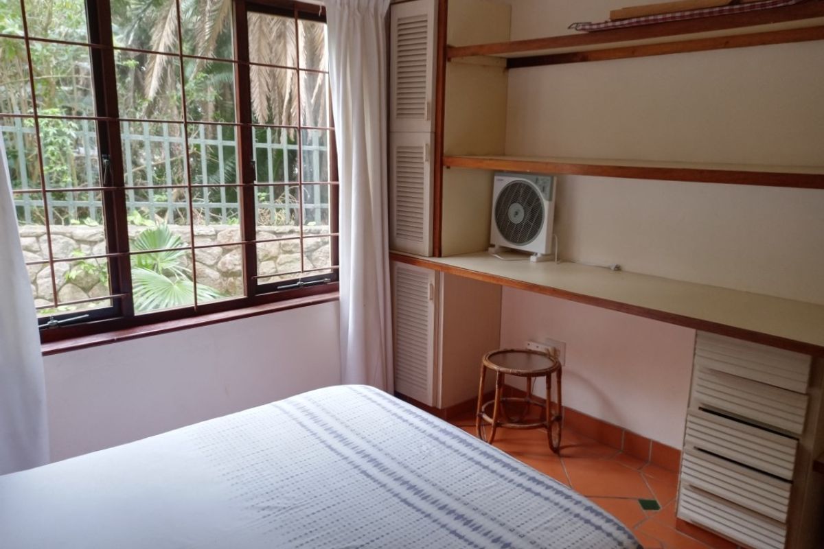 Bedroom featuring terracotta floor tiles, built-in desk with shelving, and large window with security bars overlooking the garden.