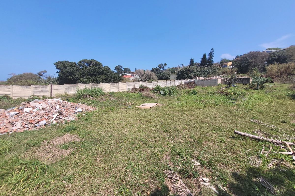 A vacant residential plot featuring a precast concrete boundary wall, overgrown grass, and a pile of construction debris.