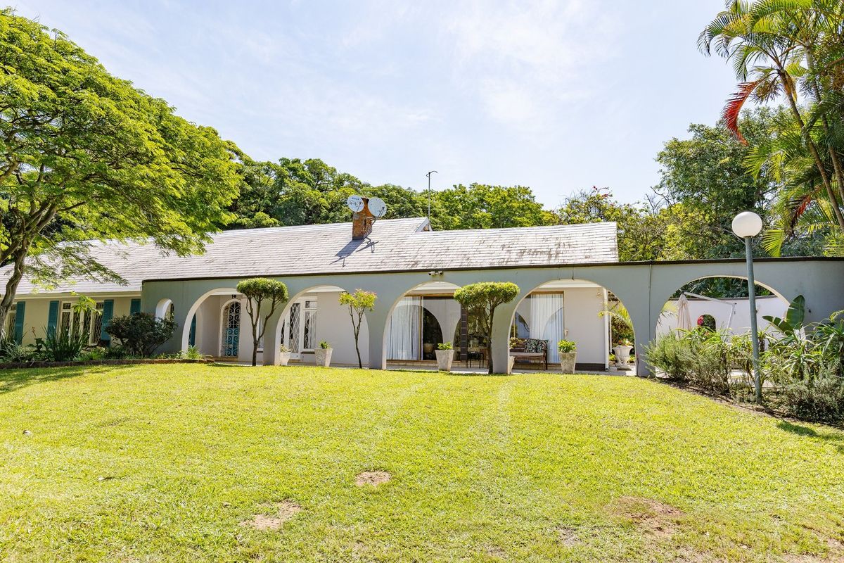 Rear view of a farm-style residence featuring a covered arched veranda, expansive lawn, and mature garden surroundings.