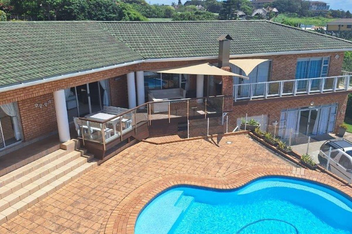 Rear exterior view featuring a brick house with a tiled roof, swimming pool, paved patio area, and a raised deck with glass balustrades.