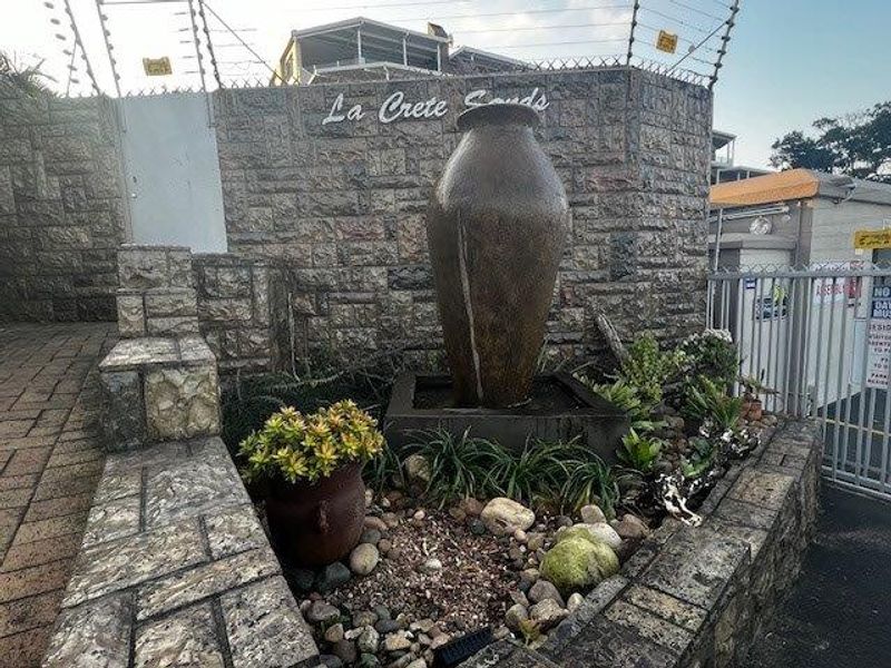 The entrance to La Crete Sands apartment complex featuring a stone-clad wall, decorative water feature, and secure gated access.