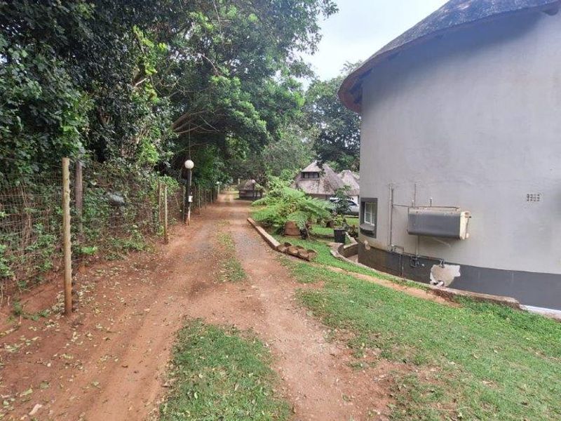 Rear exterior view of a circular townhouse featuring a thatched roof, unpaved driveway, and perimeter wire fencing.