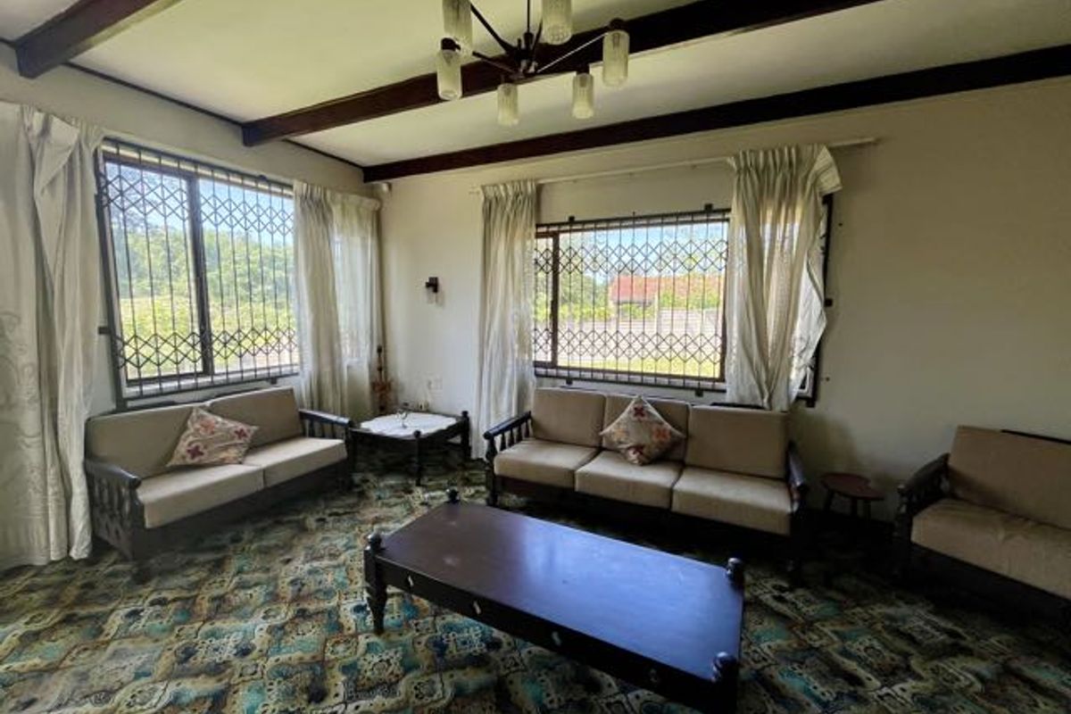 A spacious living room featuring exposed timber ceiling beams, security bars on windows, and a classic multi-arm chandelier, offering a dated but functional layout.