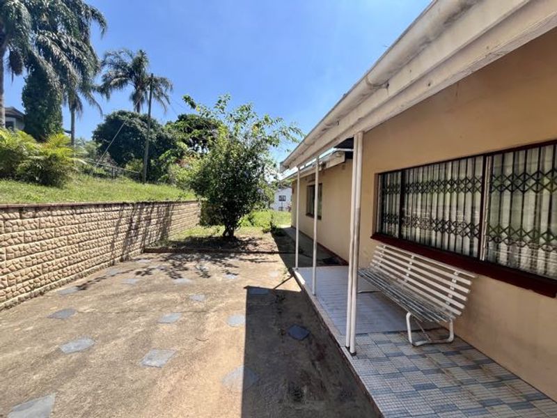 Rear exterior view featuring a covered patio area, security gates on windows, and a paved garden space with a retaining wall.