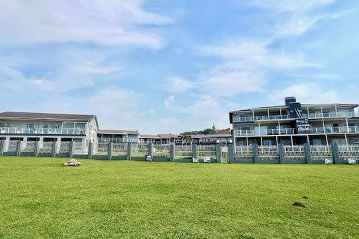 A wide exterior view of multi-story apartment buildings featuring balconies with glass balustrades, set behind a large communal lawn and perimeter fencing.