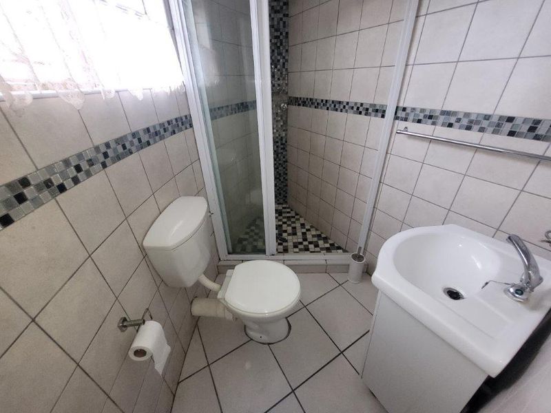 A compact bathroom featuring a corner shower enclosure, a vanity basin, and neutral wall and floor tiling with a decorative mosaic border.