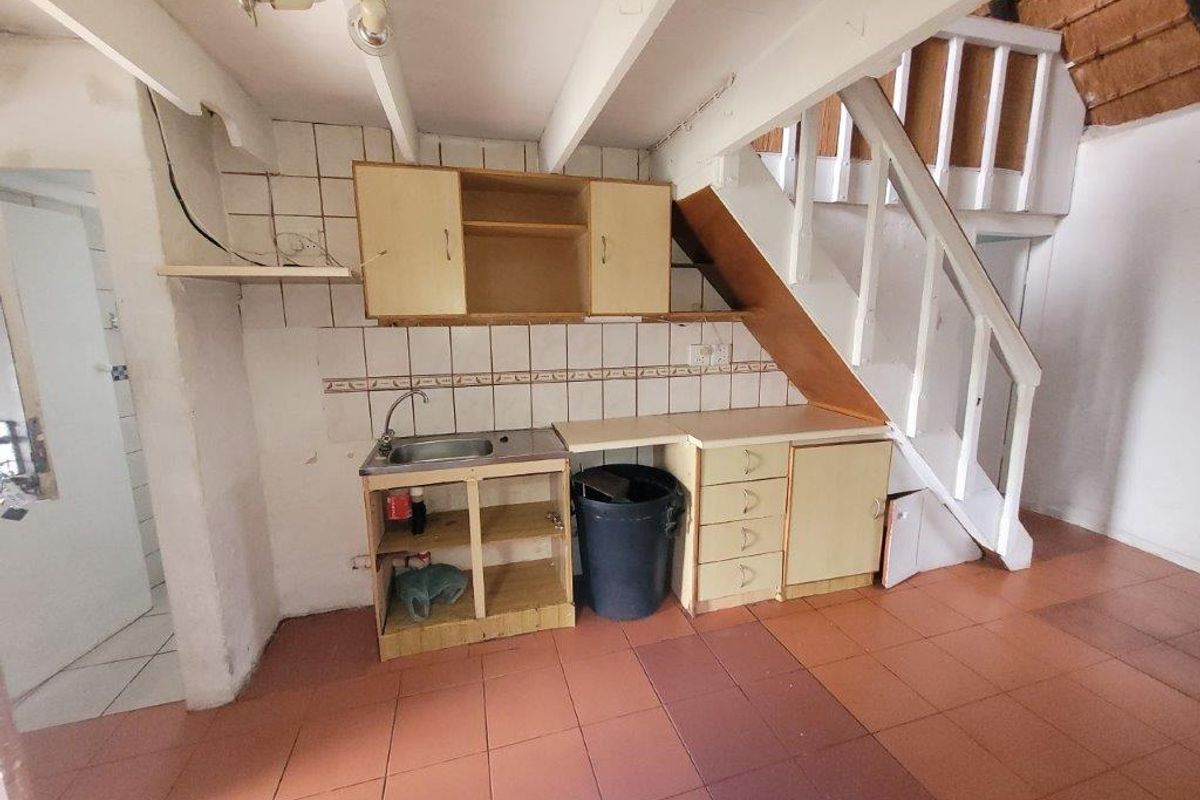A dated kitchen area featuring basic cabinetry, a sink, and tiled flooring, located beneath a staircase in a townhouse setting.