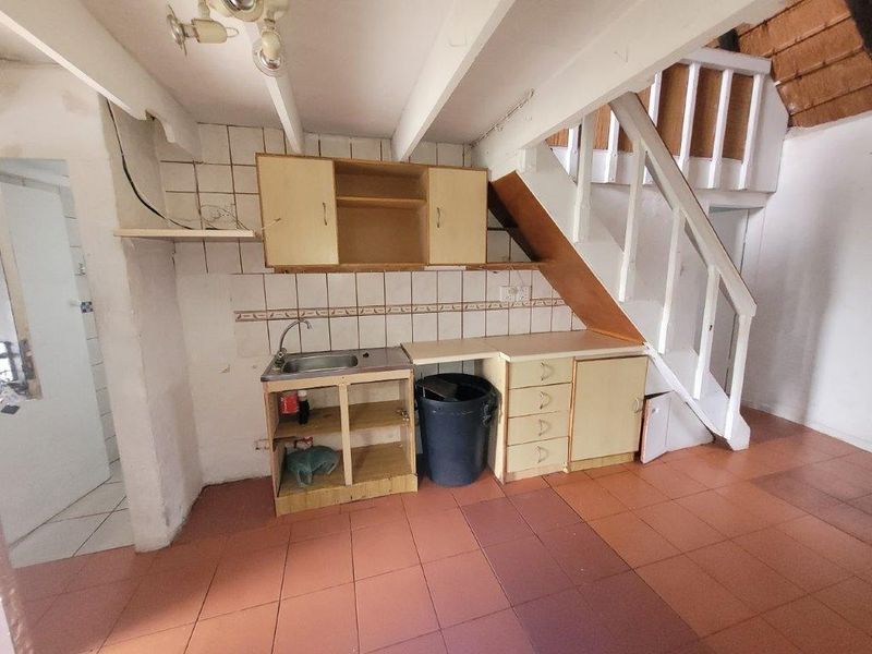 A dated kitchen area featuring basic cabinetry, a sink, and tiled flooring, located beneath a staircase in a townhouse setting.