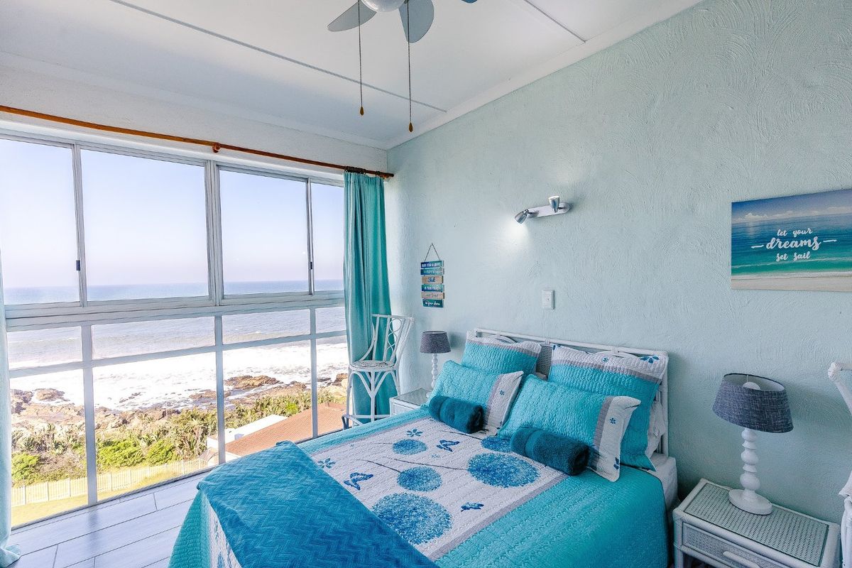 A bright bedroom featuring a large sliding window with direct ocean views, a ceiling fan, and wall-mounted lighting.