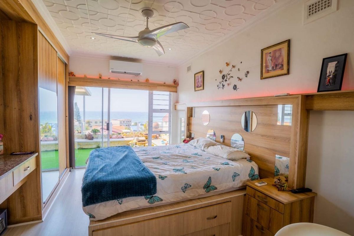 A well-appointed bedroom featuring a custom built-in headboard, mirrored wardrobes, ceiling fan, and sliding glass doors that open to a scenic coastal view.