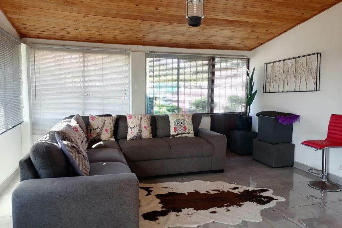 A living room featuring a timber-clad ceiling, tiled flooring, and large windows fitted with venetian blinds, providing a bright and functional living space.