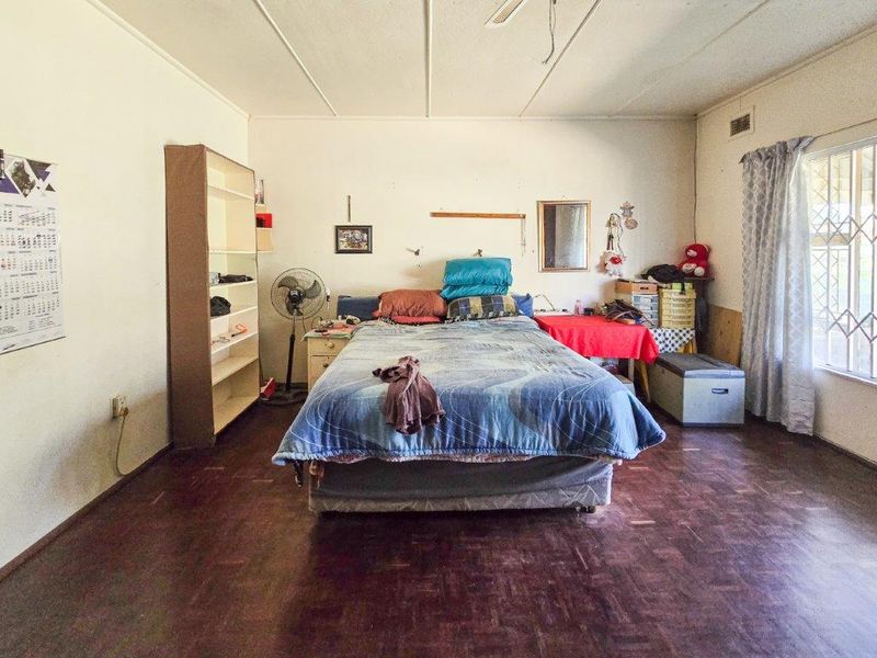 A spacious bedroom featuring parquet flooring, a ceiling fan, and a window with security bars, offering a blank canvas for renovation.