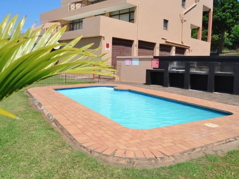 Communal swimming pool area featuring brick coping, a built-in braai facility, and landscaped lawn, set against the apartment building exterior.