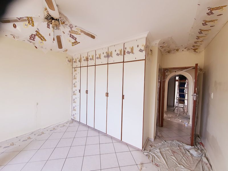 A bedroom currently undergoing renovations, featuring built-in cupboards, tiled flooring, and a ceiling fan, with protective plastic sheeting still in place.