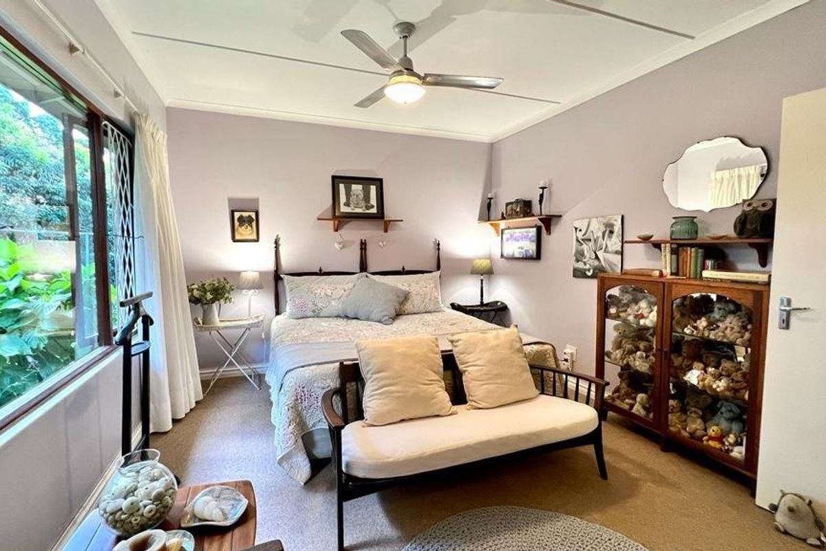 A well-lit bedroom featuring a ceiling fan, large window with garden views, and neutral carpeting.