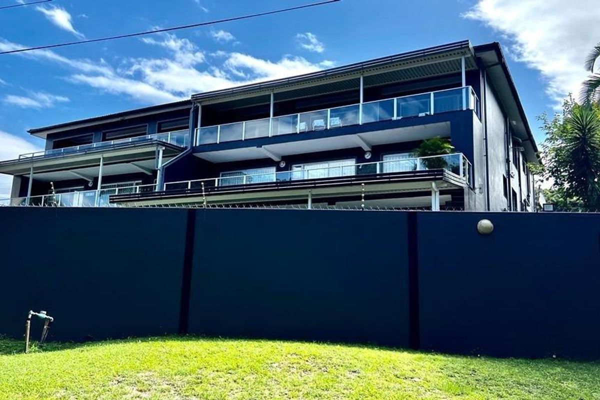 Rear view of a multi-story apartment building featuring private balconies with glass balustrades, enclosed by a secure perimeter wall with electric fencing.