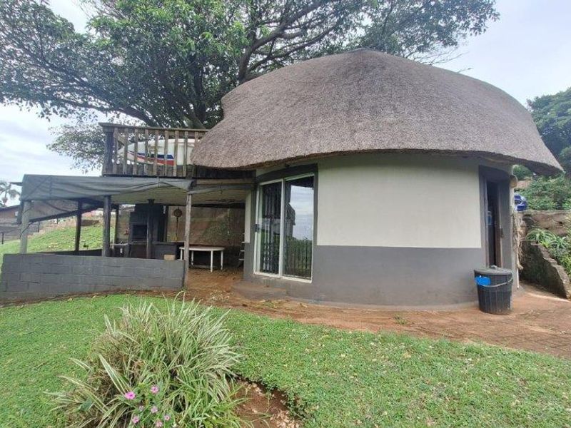 A unique circular townhouse unit featuring a traditional thatched roof, an attached covered patio area, and a raised wooden deck nestled under mature trees.