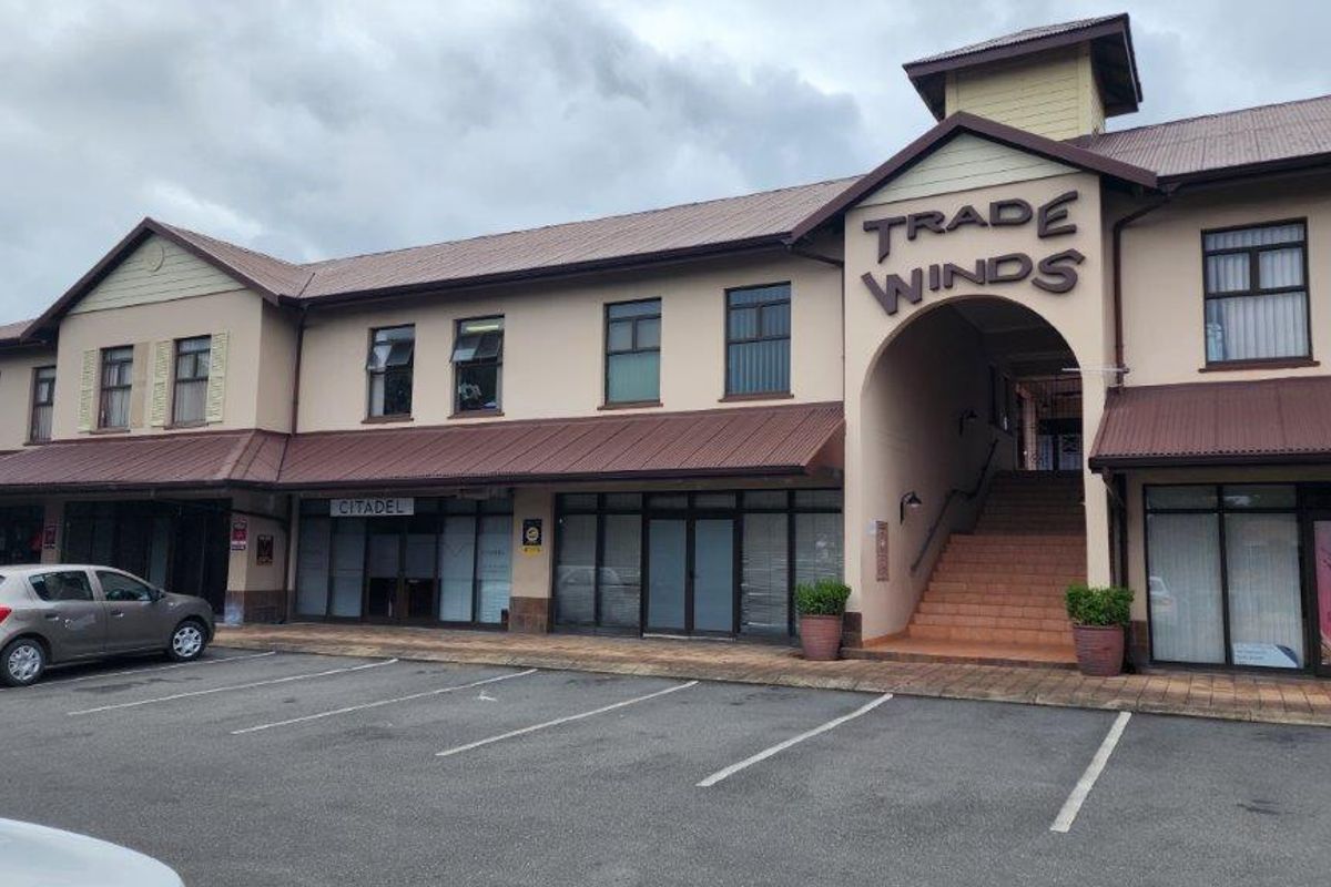 Front elevation of the Trade Winds commercial building featuring a paved parking area, multiple glass storefronts, and a central staircase entrance.