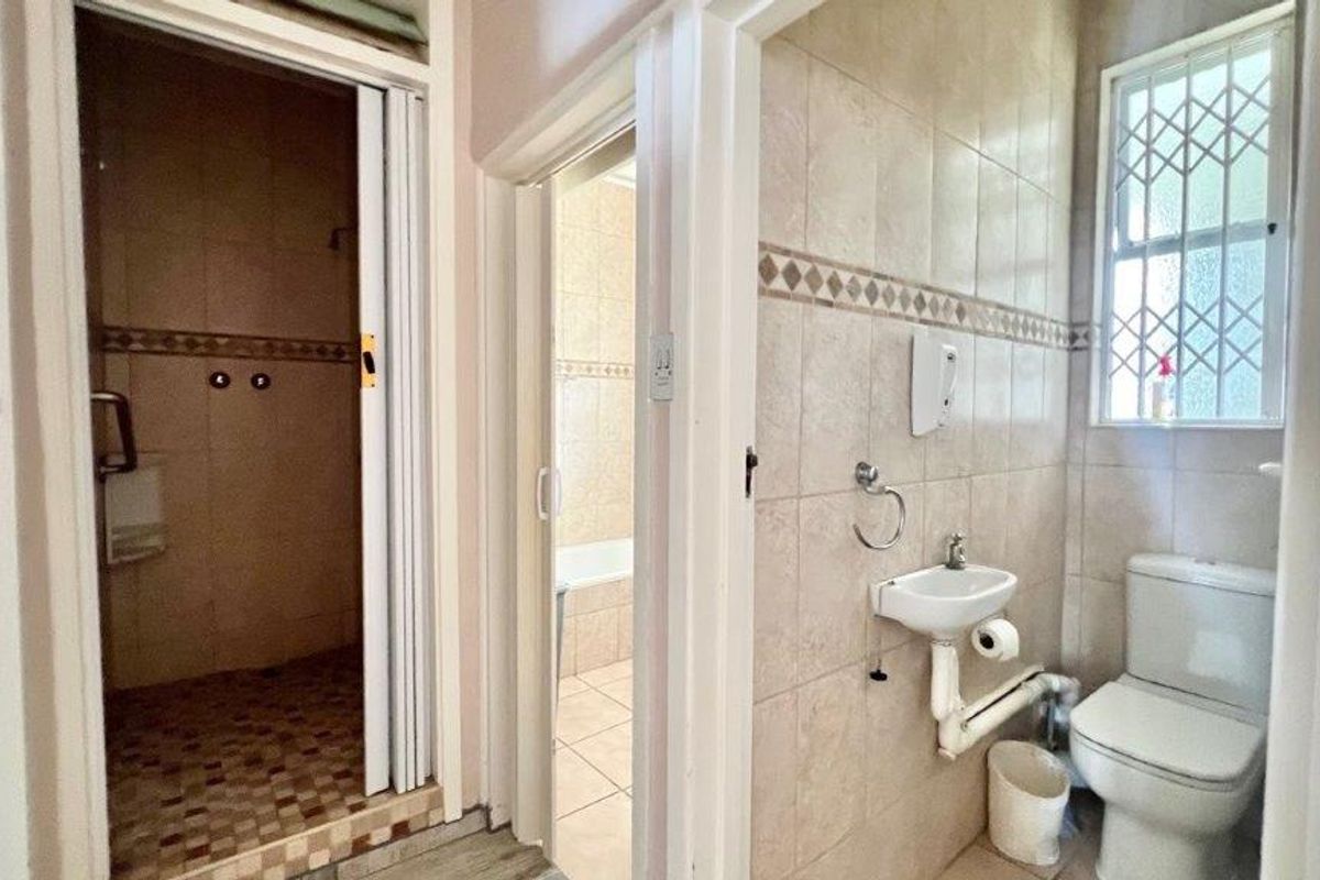A functional bathroom featuring a toilet, wall-mounted basin, and a separate shower cubicle with tiled finishes and security bars on the window.