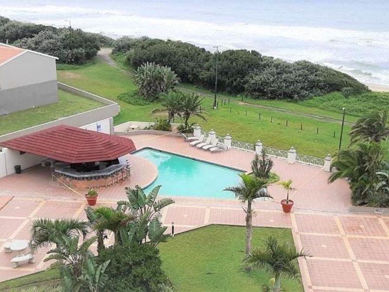 Aerial view of a communal pool area featuring a covered bar, built-in braai, paved patio, and landscaped gardens with direct beach access.