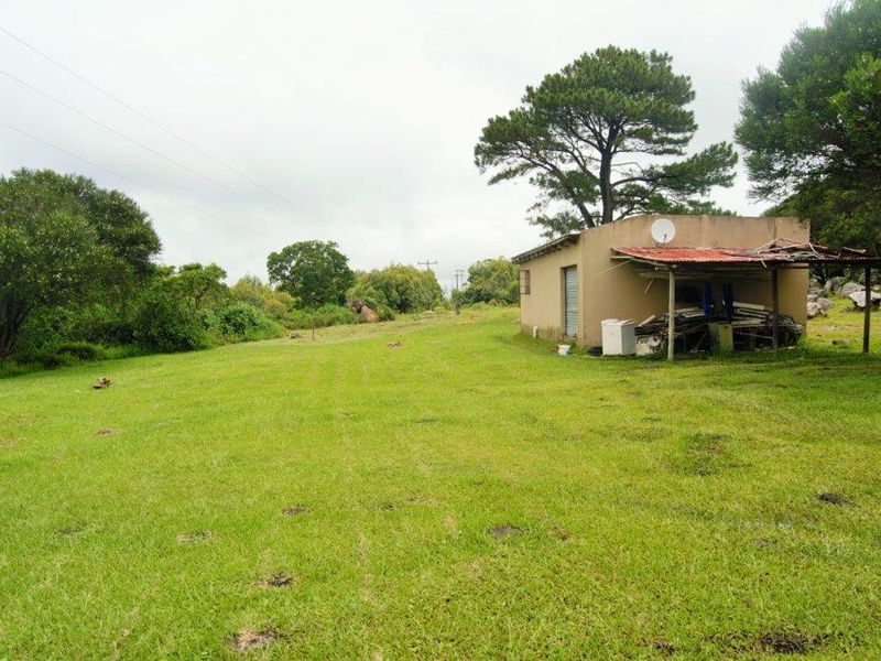 A view of a small outbuilding on a large grassy farm plot, featuring a weathered structure with a damaged roof canopy.