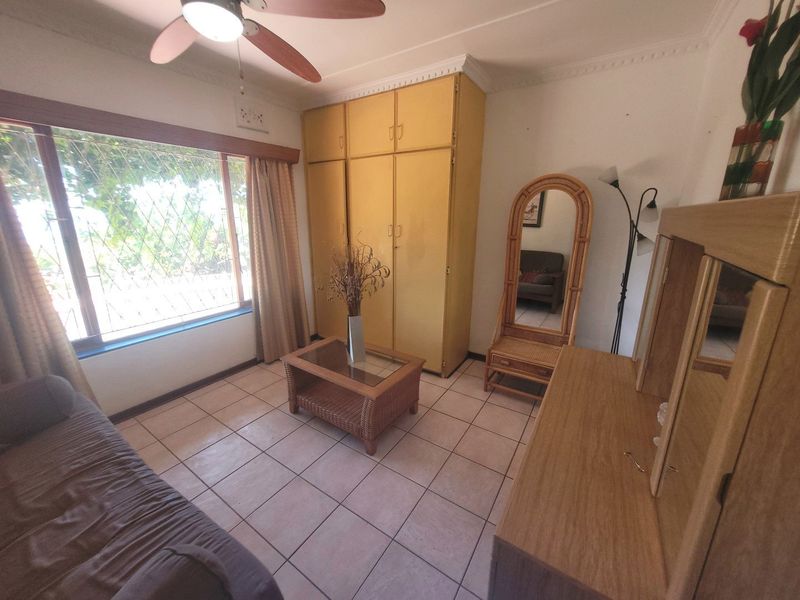 A bedroom featuring tiled flooring, built-in cupboards, and a ceiling fan, with security bars installed on the window.
