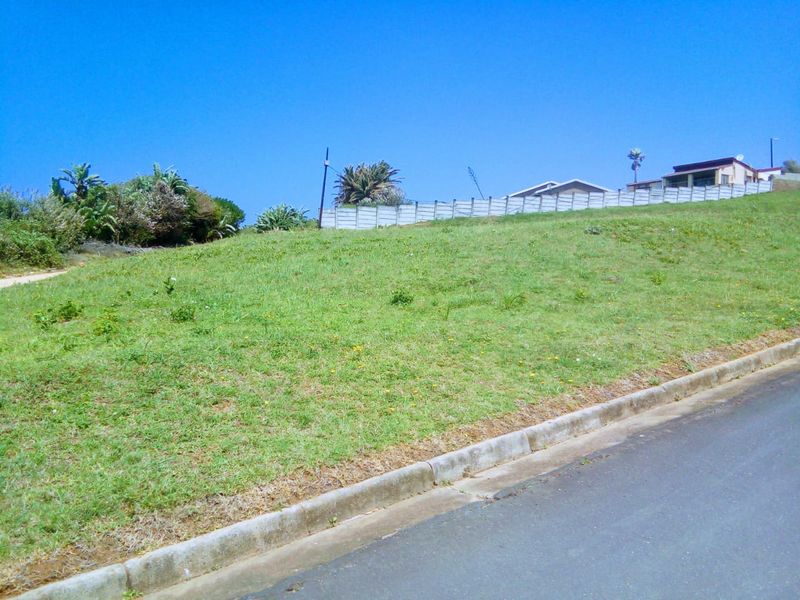 A vacant residential plot featuring a gentle slope, grass cover, and concrete boundary walling, situated along a paved street.