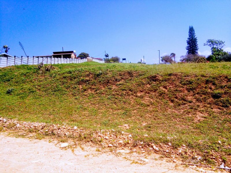 A view of a vacant residential plot featuring a sloped grassy area and perimeter fencing, situated along an unpaved road.