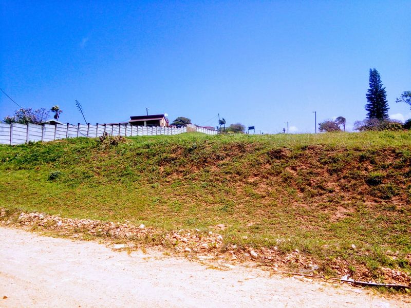 A view of a vacant residential plot featuring a sloped grassy area, perimeter walling, and access from an unpaved road.