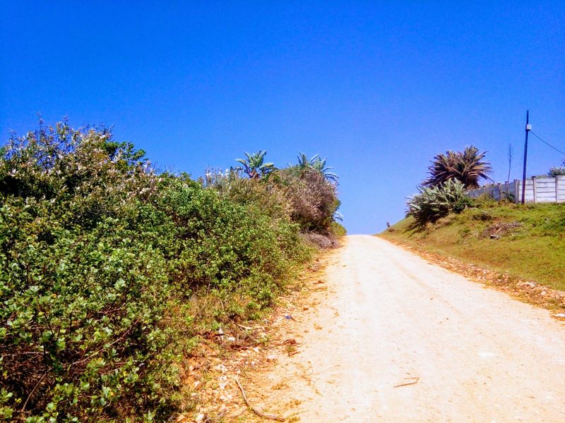 A view of an unpaved dirt access road leading up a slope, bordered by natural vegetation and a boundary wall.