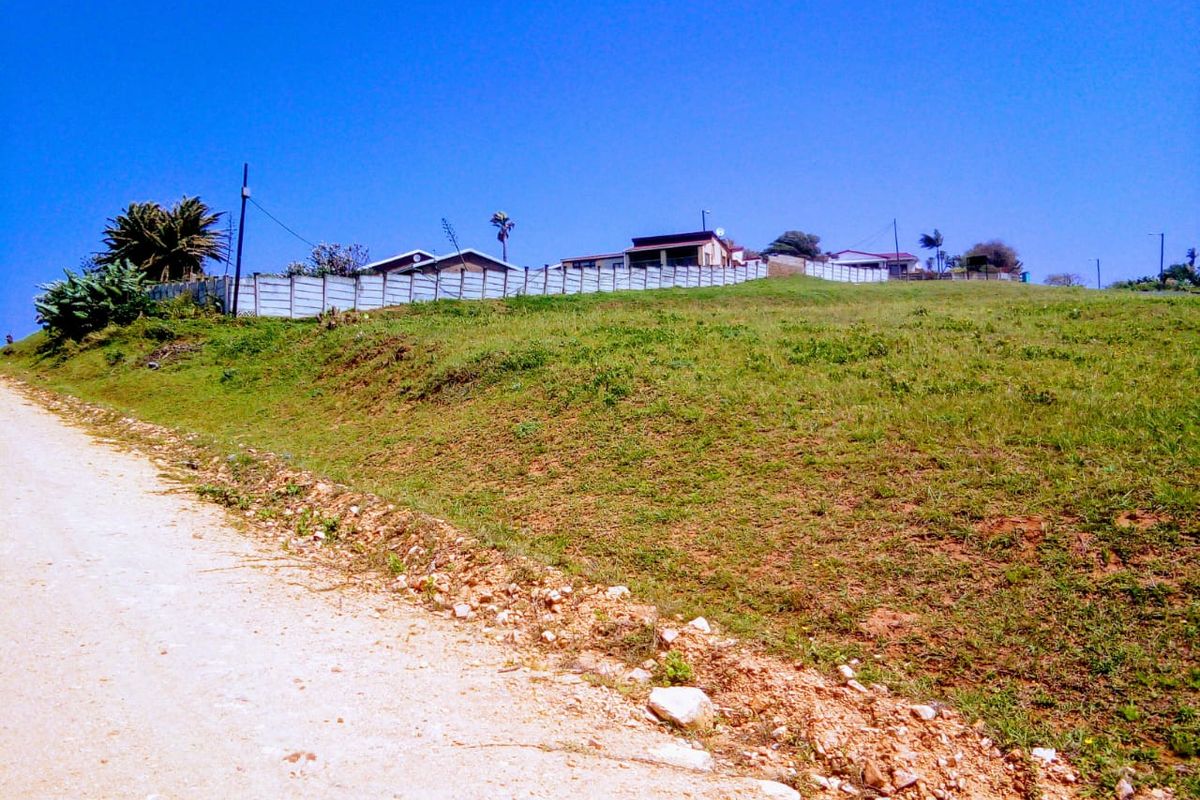 A view of a vacant residential plot from the adjacent dirt road, featuring a gentle slope and perimeter fencing.