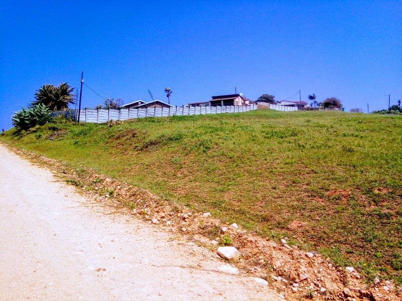 A view of a vacant residential plot from the adjacent dirt road, featuring a gentle slope and perimeter fencing.