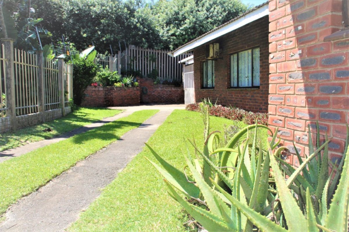 A view of the townhouse exterior featuring a brick facade, concrete driveway, and established garden area with perimeter fencing.