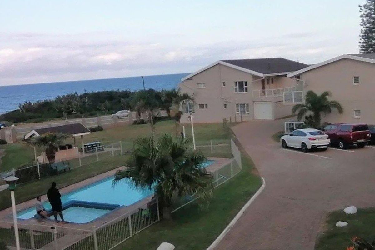 Elevated view of the apartment complex grounds, featuring a swimming pool, paved driveway, and coastal scenery.