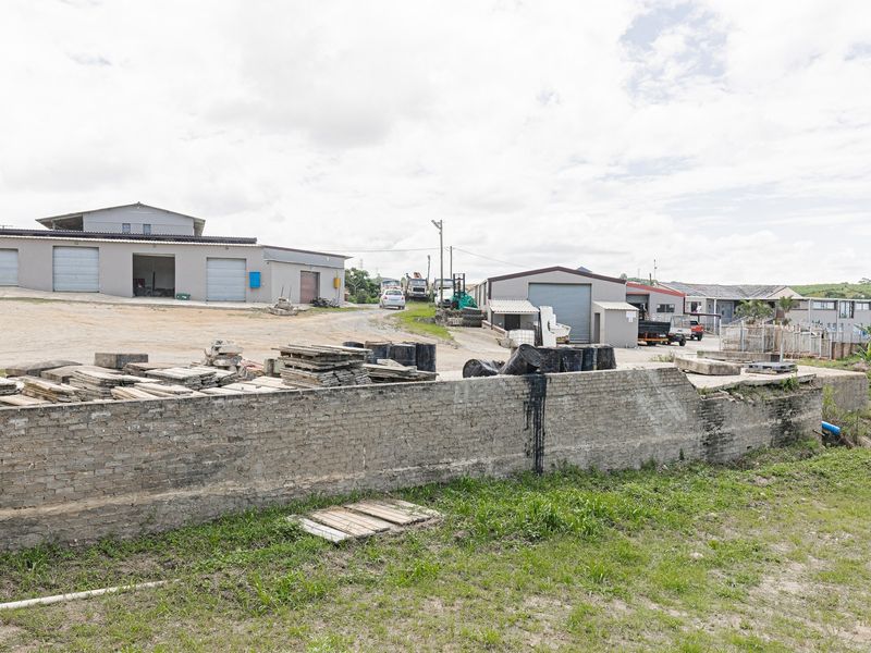 Rear view of an industrial property featuring multiple warehouse units with roller shutter doors, a large unpaved yard, and a retaining wall.
