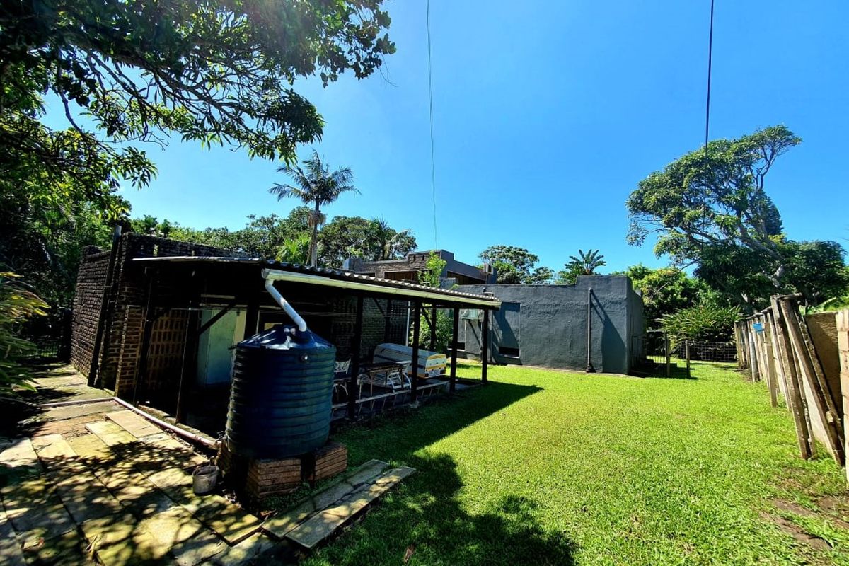 Rear exterior view of the property featuring a covered patio area, water tank, paved walkway, and a spacious lawn with boundary fencing.