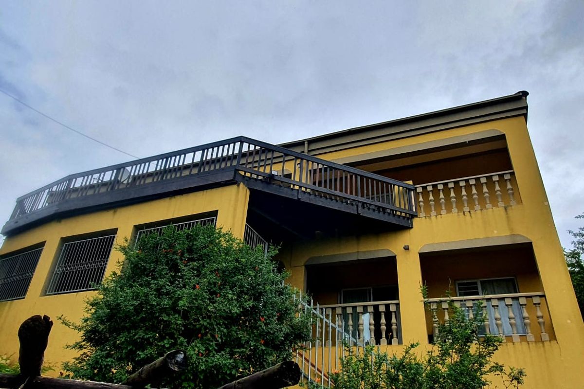 A low-angle view of a two-story yellow house featuring a large upper-level wooden balcony and lower-level patio with decorative balustrades.