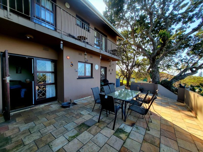 A spacious rear patio featuring stone paving, accessible via sliding doors, with an overhead balcony and mature trees providing shade.