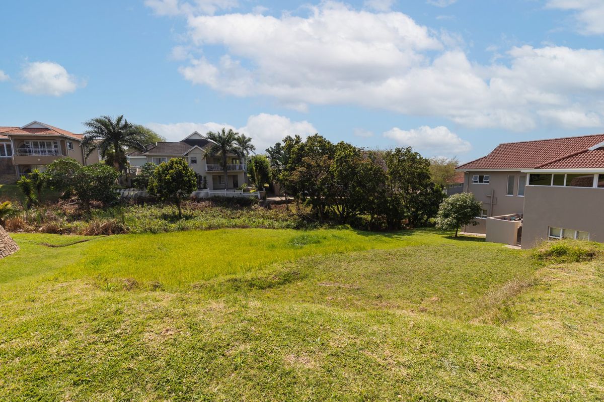 A view of a vacant residential plot featuring established grass, surrounding mature trees, and neighboring residential properties in a quiet suburban setting.